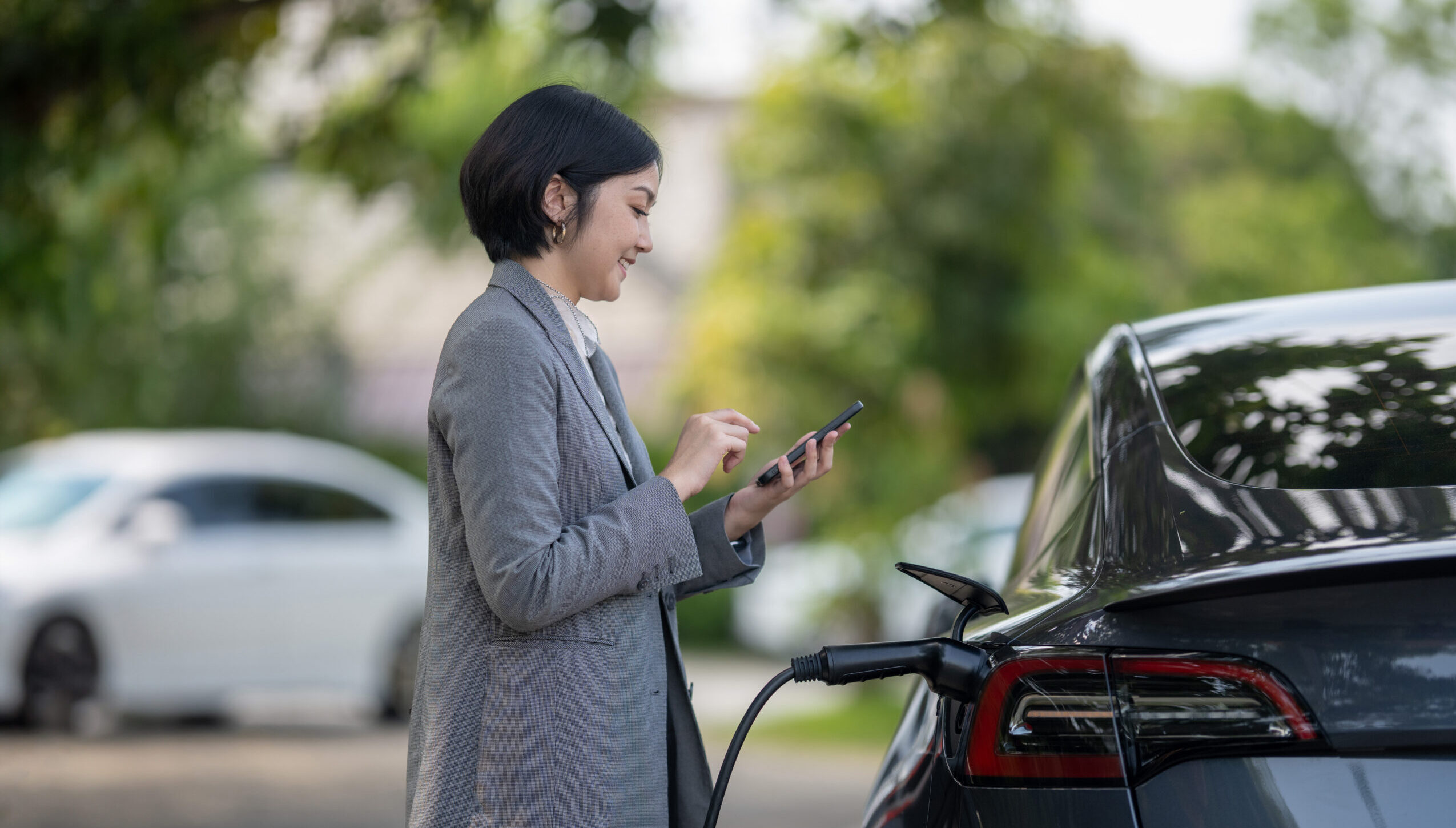 Progressive businesswoman using smartphone learning and check the charging time of electric car at charging station. Eco friendly rechargeable EV car concept.